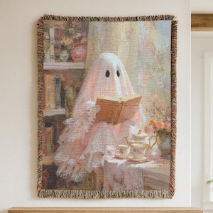 May include: Woven tapestry featuring a ghost reading a book while enjoying tea. The ghost is draped in a pink and white fabric with lace trim. A tea set and flowers are on a table in front of the ghost. The tapestry has a fringed border.