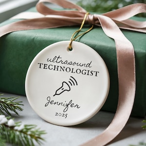 Könnte beinhalten: Runder, weißer Keramikschmuck mit dem Text "ultrasound TECHNOLOGIST" und dem Namen "Jennifer" unter einer Sonogramm-Grafik. Der Schmuck hängt an einer goldenen Schnur und liegt auf einer verpackten grünen Geschenkbox mit einem rosa Band.