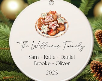 May include: A round, cream-colored Christmas ornament with a watercolor illustration of cookies on a plate. The text "The Williams Family" is above the names Sam, Katie, Daniel, Brooke, and Oliver, with the year 2023 below.