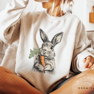 May include: Cream-colored sweatshirt featuring a detailed illustration of a rabbit holding a carrot. The rabbit is depicted in shades of gray, with the carrot in orange and green. The sweatshirt has long sleeves and a crew neck.
