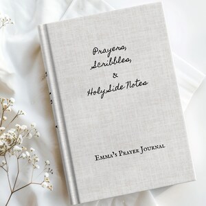 Puede incluir: Un diario de color claro con el texto "Prayers, Scribbles, & HolySide Notes" y "Emma's Prayer Journal" impreso en la portada. El diario está sobre una tela blanca, con flores secas en primer plano.