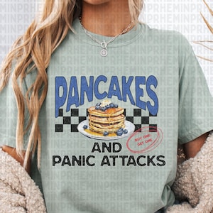May include: A light green t-shirt with the text "Pancakes and Panic Attacks" in black and blue lettering. The text is surrounded by a checkered pattern and a graphic of a stack of pancakes with blueberries and syrup.