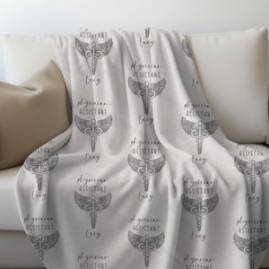 Könnte beinhalten: Weiße Fleece-Decke mit einem sich wiederholenden Muster aus einem medizinischen Caduceus-Symbol mit Flügeln und dem Text "Arztassistent" und dem Namen "Lucy".