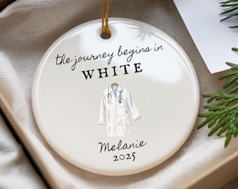 Könnte beinhalten: Runder weißer Keramik-Weihnachtsschmuck mit goldenem Aufhänger. Er zeigt eine Illustration eines Arztmantels und Stethoskops mit dem Text "the journey begins in WHITE", dem Namen "Melanie" und dem Jahr "2025".