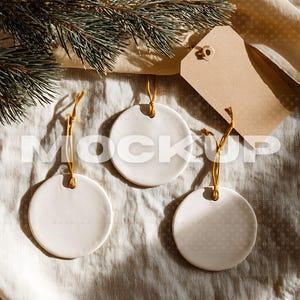 Könnte beinhalten: Drei weiße Keramik-Ornamente mit goldenen Schnüren, angeordnet auf einer strukturierten Oberfläche. Ein braunes Etikett und ein Tannenzweig befinden sich im Hintergrund. Die Ornamente haben eine runde Form und sind bereit zur Individualisierung.
