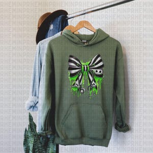 Op de afbeelding: Olijfgroene hoodie met een zwart-wit gestreept strikdesign, met schedels en groene druipaccenten. De hoodie heeft een voorzak en een capuchon met trekkoord. Het ontwerp bevat een stethoscoop.