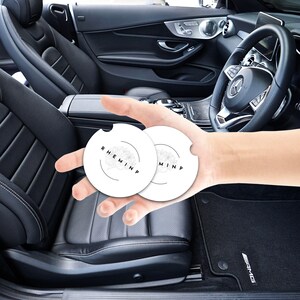 May include: Two white car coasters with a black floral design and the word "RHEMINP" printed on them. The coasters are held in front of a black leather car interior, including the steering wheel and seats.