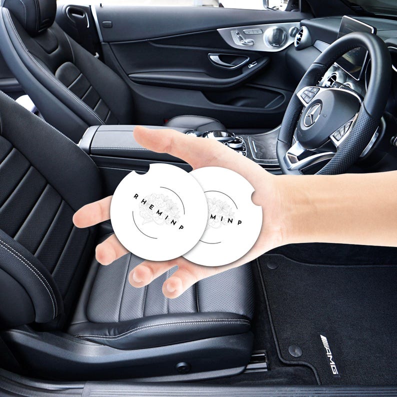 May include: Two white car coasters with a black floral design and the word "RHEMINP" printed on them. The coasters are held in front of a black leather car interior, including the steering wheel and seats.