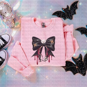 May include: A light pink sweatshirt featuring a large black bow with a nurse's cap and bandages. Red drips and a stethoscope are also part of the design. Black bat decorations and a disco ball skull are in the background.