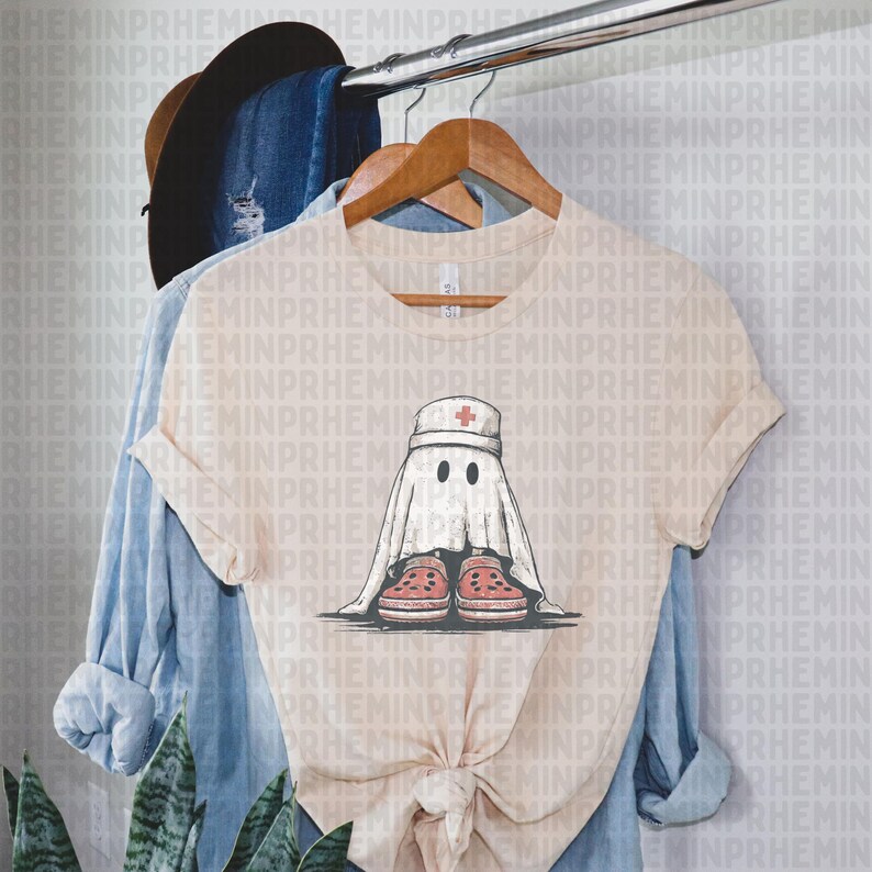 Pode incluir: Camiseta bege com um fantasma de desenho animado usando um chap&eacute;u de enfermeira e tamancos vermelhos. O fantasma tem dois olhos pretos e est&aacute; envolto em um len&ccedil;ol branco. Uma camisa de manga comprida azul clara e um chap&eacute;u marrom pendurados em um cabide.