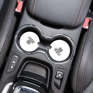 May include: Two white car coasters with black text that reads "CUSTOM HERE" in a car's cup holders. The coasters are round and appear to be made of absorbent material. The car interior is black with red stitching.