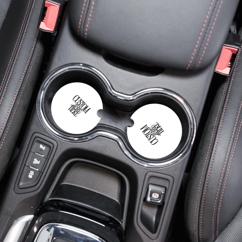 May include: Two white car coasters with black text that reads "CUSTOM HERE" in a car's cup holders. The coasters are round and appear to be made of absorbent material. The car interior is black with red stitching.