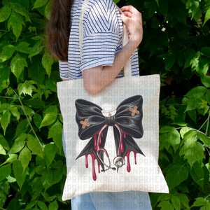 May include: A cream-colored tote bag featuring a large black bow with a nurse's hat and bandages. Red blood-like drips and a stethoscope are below the bow. The bag is held by a person wearing a striped shirt.