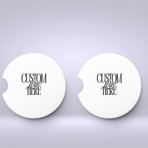 May include: Two white car coasters with a curved notch and the text "CUSTOM Logo HERE" in black script. The coasters are round and sit on a light purple background.