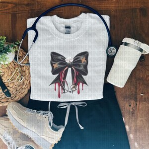 May include: White crew neck t-shirt with a black bow design featuring a nurse's cap, bandages, and a stethoscope. The design includes red drips. A blue stethoscope, black scrubs, and a white tumbler are also visible.