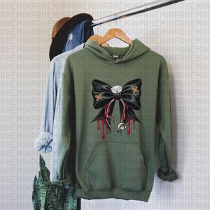 May include: Olive green hoodie with a black bow design featuring a nurse's cap, bandages, and a stethoscope with dripping blood. The hoodie has a front pocket and is hanging on a wooden hanger.