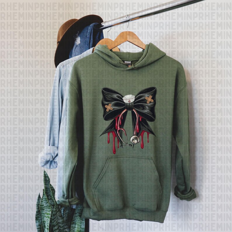 May include: Olive green hoodie with a black bow design featuring a nurse's cap, bandages, and a stethoscope with dripping blood. The hoodie has a front pocket and is hanging on a wooden hanger.