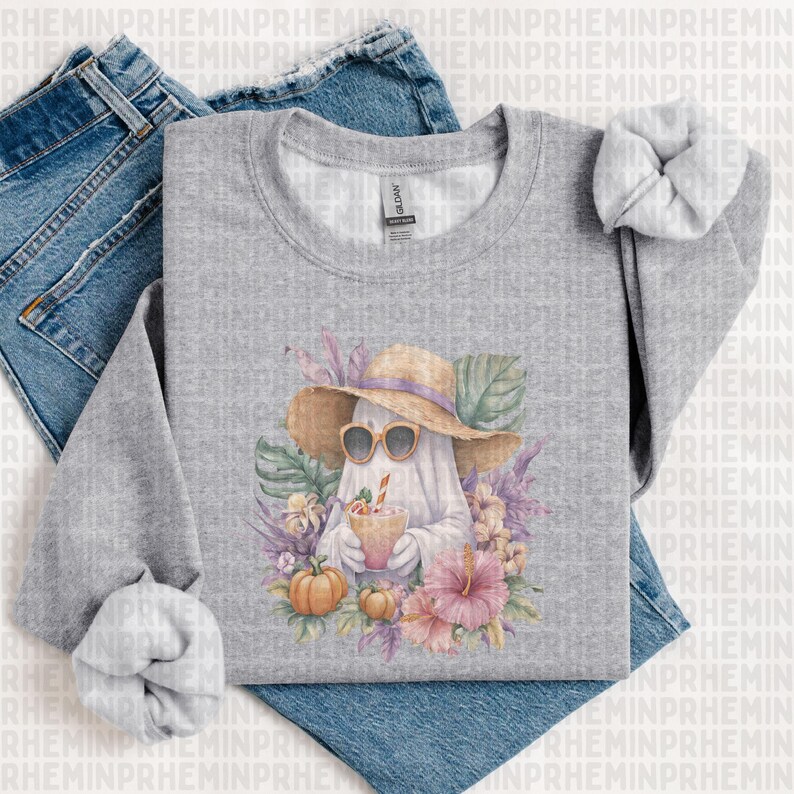 May include: A gray sweatshirt featuring a watercolor illustration of a ghost wearing sunglasses and a straw hat, holding a drink. The design includes pumpkins, flowers, and tropical leaves. The sweatshirt is displayed on top of blue jeans.