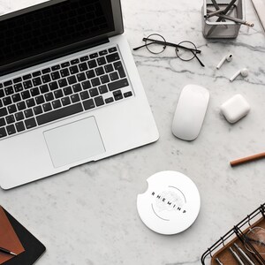 May include: A top-down view of a workspace with a laptop, mouse, and wireless earbuds. A white car coaster with the text "RHEMINP" is in the center. Other items include a notebook, pen, glasses, and a small succulent plant.