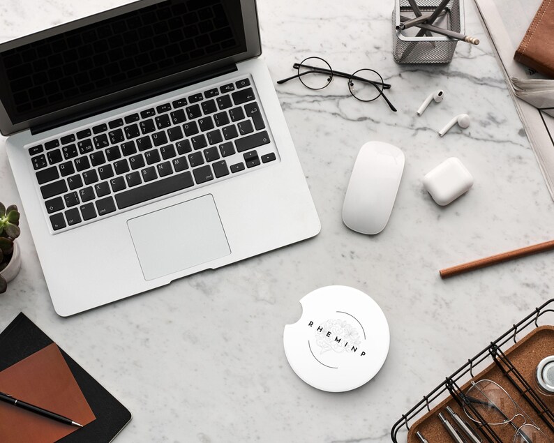 May include: A top-down view of a workspace with a laptop, mouse, and wireless earbuds. A white car coaster with the text "RHEMINP" is in the center. Other items include a notebook, pen, glasses, and a small succulent plant.