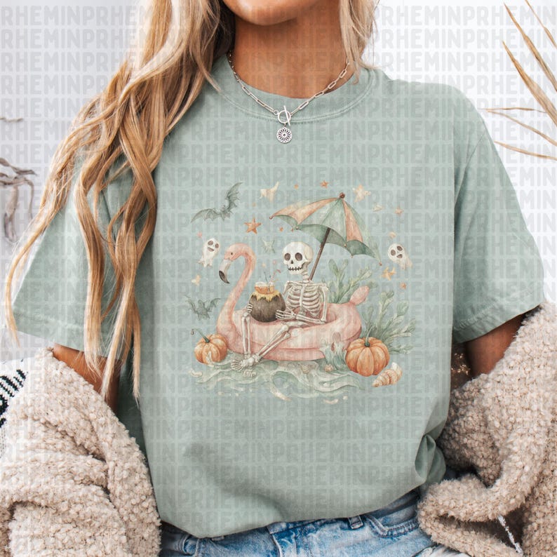 Op de afbeelding: Lichtgroen T-shirt met een Halloween-thema. Het toont een skelet op een roze flamingo-vlot, omringd door pompoenen, vleermuizen en geesten. Het skelet staat onder een kleine parasol.