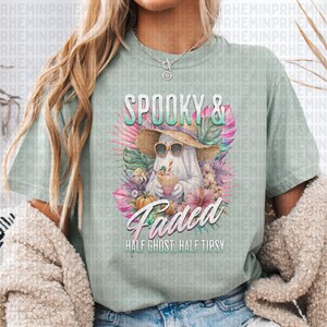 May include: A light green t-shirt with a graphic featuring a ghost wearing sunglasses and a straw hat, holding a drink. The text reads "Spooky & Faded, Half Ghost, Half Tipsy". The design includes tropical flowers and pumpkins.