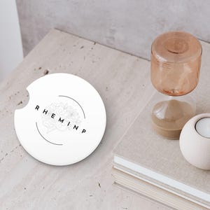 May include: A white car coaster with a floral design and the word "RHEMINP" in black lettering. The coaster is next to a small hourglass and a white candle holder with a lit candle.