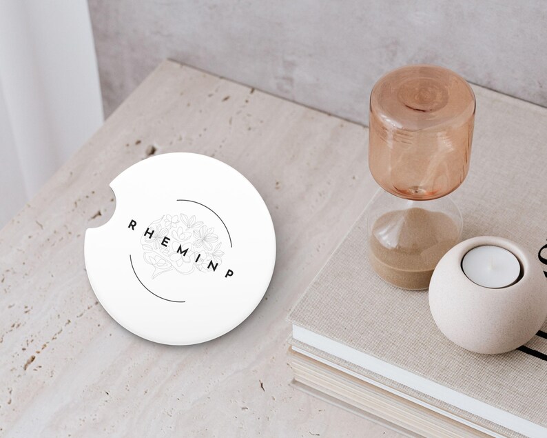 May include: A white car coaster with a floral design and the word "RHEMINP" in black lettering. The coaster is next to a small hourglass and a white candle holder with a lit candle.