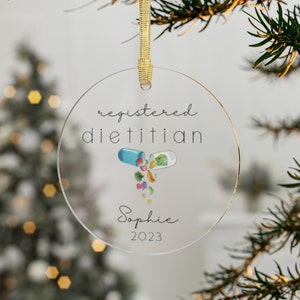 May include: A clear round Christmas ornament with the text "registered dietitian" and a graphic of a pill with healthy food spilling out. The ornament also includes the name "Sophie" and the year "2023".