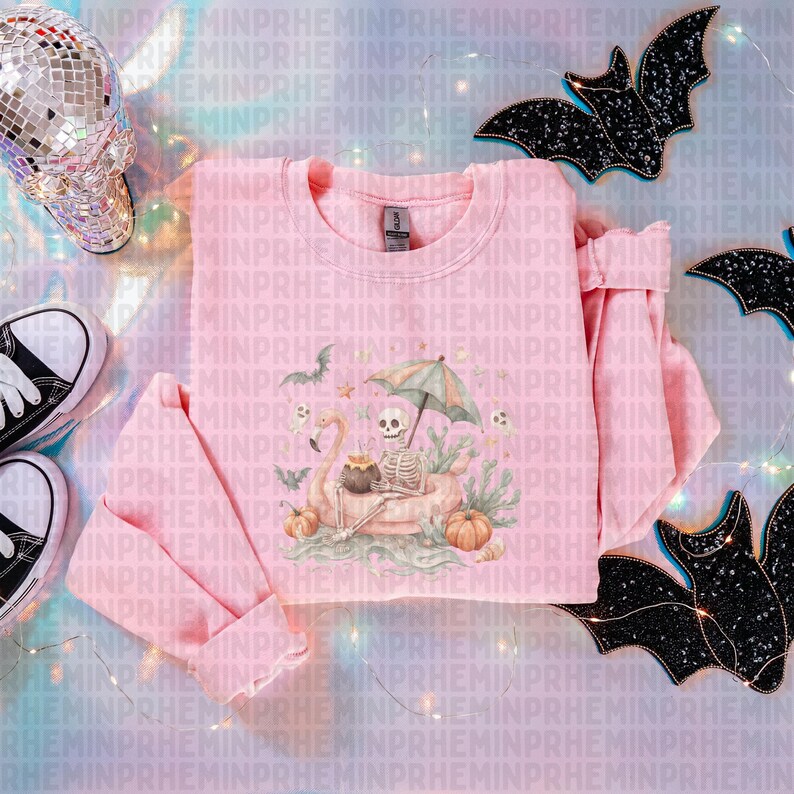 Op de afbeelding: Een lichtroze sweatshirt met een skelet op een flamingo-vlot, pompoenen en vleermuizen. Het ontwerp is gedrukt op een roze sweatshirt. Zwart-witte sneakers en een discobal schedel staan op de achtergrond.