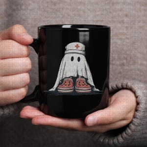 Pode incluir: Caneca de cer&acirc;mica preta com um desenho de fantasma de desenho animado. O fantasma usa um chap&eacute;u de enfermeira com uma cruz vermelha e tamancos vermelhos. O fantasma est&aacute; envolto em um len&ccedil;ol branco e tem dois olhos pretos. A caneca &eacute; segurada por uma pessoa.