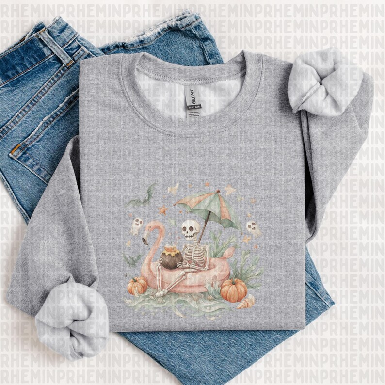 Op de afbeelding: Een grijze sweatshirt met een skelet dat ontspant op een roze flamingo-vlot onder een paraplu. Het ontwerp bevat pompoenen, geesten en andere Halloween-elementen. De sweatshirt wordt gecombineerd met een blauwe jeans en een scrunchie.