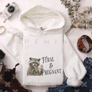 Op de afbeelding: Witte hoodie met een wasbeer die een augurk en ijs eet. De tekst "Feral & Pregnant" staat op de hoodie gedrukt.