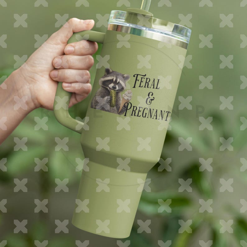 Op de afbeelding: Een groene reistumbler van roestvrij staal met een zwarte handgreep en een wasbeer-afbeelding. Op de tumbler staat de tekst "Feral & Pregnant".