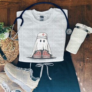 Pode incluir: Camisola branca de gola redonda com um desenho de fantasma usando um chap&eacute;u de enfermeira e tamancos vermelhos. Um estetosc&oacute;pio e um copo branco s&atilde;o colocados perto da camisa. A camisa &eacute; combinada com um uniforme preto e t&ecirc;nis bege.