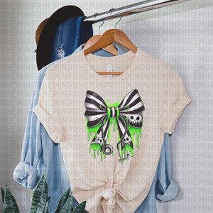 Op de afbeelding: Beige T-shirt met een zwart-wit gestreept strikdesign, groene druppelaccenten en schedeldetails. Een stethoscoop is ook inbegrepen. Het shirt hangt aan een houten hanger.