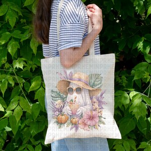 May include: A beige tote bag featuring a watercolor illustration of a ghost wearing sunglasses and a straw hat, holding a drink. The design includes pumpkins, flowers, and tropical leaves. The bag is held by a person.