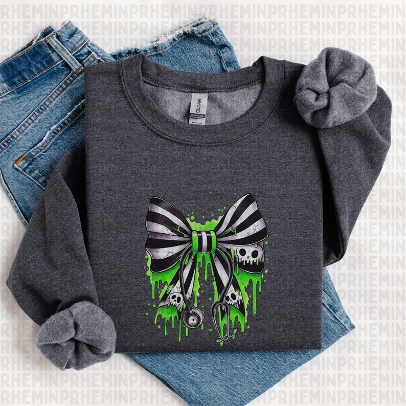 Op de afbeelding: Donkergrijze sweatshirt met een zwart-wit gestreepte strik, een groen midden en druipend groen slijm. De strik heeft schedelaccenten en een stethoscoopontwerp. De sweatshirt wordt gepresenteerd op een blauwe jeans.