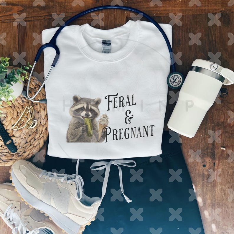Op de afbeelding: Wit sweatshirt met een zwarte wasbeer afbeelding en de tekst "Feral & Pregnant". Het sweatshirt ligt op een houten oppervlak met een paar witte sneakers, een zwarte broek en een witte waterfles.