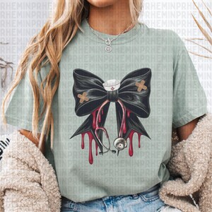 May include: A light green t-shirt featuring a large black bow with a nurse's hat and a stethoscope. The bow has two bandages and red drips. The shirt is a comfortable, casual style.