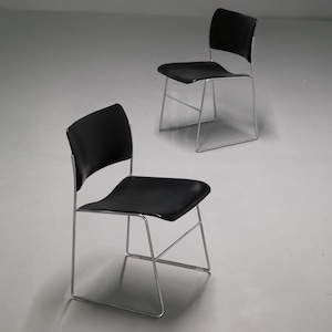 May include: Two modern black chairs with curved backs and seats. The chairs have a minimalist design with a sleek, black seat and backrest supported by a silver metal frame. The chairs are set against a neutral background.