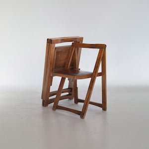 May include: A set of three wooden folding chairs. The chairs are made of a light brown wood and have a simple, classic design. One chair is unfolded, while the other two are folded and stacked together. The chairs are suitable for indoor or outdoor use.