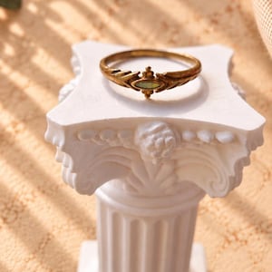 Könnte beinhalten: Ein goldfarbener Metallring mit einem zentralen, marquiseförmigen hellgrünen Stein. Die Ringschiene weist auf beiden Seiten der Steinfassung ein strukturiertes, blattartiges Design auf. Der Ring liegt auf einer kleinen, weißen Säule.