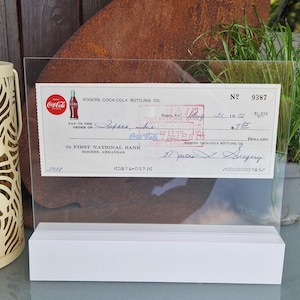 Puede incluir: Un cheque bancario antiguo de Rogers Coca-Cola Bottling Co. en un soporte de exhibición de acrílico transparente con base blanca. El cheque muestra el logotipo de Coca-Cola, la fecha 31 de agosto de 1972, un sello de pagado y una firma.