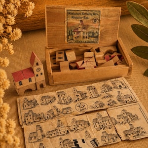 Könnte beinhalten: Eine Holzkiste mit der Aufschrift Miniature Building Box, Miniatur-Bau, die verschiedene kleine Holzklötze, ein kleines zusammengebautes Holzgebäude mit rotem Dach und ein gefaltetes Papierblatt mit Abbildungen von Gebäuden enthält.