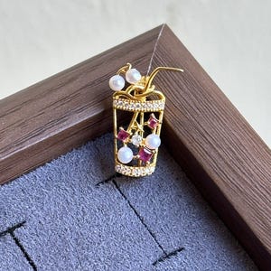 May include: A gold-colored pendant shaped like a beverage cup, adorned with white pearls and red gemstones. The cup features a straw and is set on a gray surface within a wooden frame. The pendant is likely a piece of jewelry.