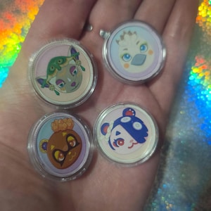 May include: Four circular acrylic charms featuring colorful cartoon character faces. Each charm is encased in a clear protective plastic shell. The designs include a green creature, a white bird-like figure, a brown bear, and a blue and white bear.