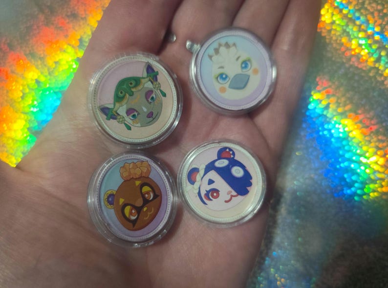 May include: Four circular acrylic charms featuring colorful cartoon character faces. Each charm is encased in a clear protective plastic shell. The designs include a green creature, a white bird-like figure, a brown bear, and a blue and white bear.