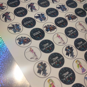 May include: A sheet of circular stickers featuring various characters and text from the Legend of Zelda series. The designs include illustrations of Urbosa, Mipha, Revali, Daruk, and Zelda, with text labels such as Zelda Super Smash Bros and Breath of the Wild.