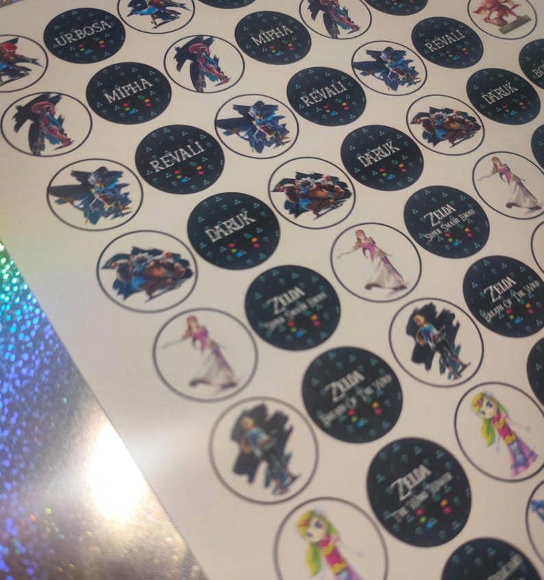May include: A sheet of circular stickers featuring various characters and text from the Legend of Zelda series. The designs include illustrations of Urbosa, Mipha, Revali, Daruk, and Zelda, with text labels such as Zelda Super Smash Bros and Breath of the Wild.